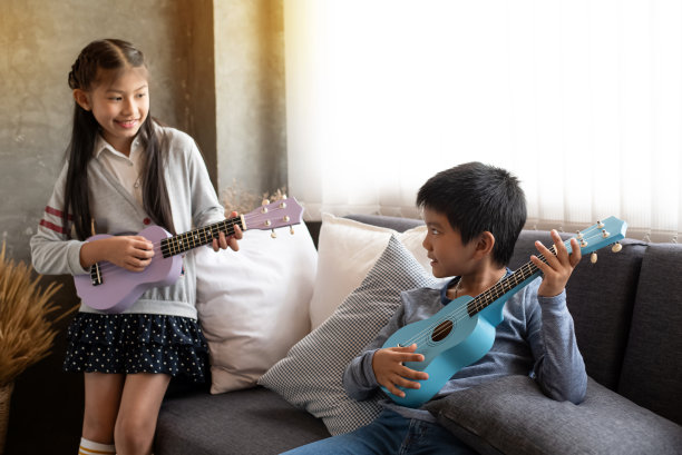 在选择性的聚焦下，小男孩一边弹着四弦琴，一边对着模糊的可爱女孩微笑，坐在沙发的左边，在家里的工作室里，模糊的灯光环绕图片下载