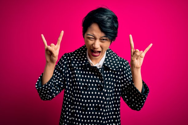 年轻漂亮的亚洲女孩穿着休闲夹克，在粉红色背景前做出摇滚手势，表达疯狂图片下载