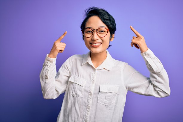年轻美丽的亚洲女孩穿着休闲衬衫和眼镜站在紫色的背景微笑指着头与双手手指，伟大的想法或想法，良好的记忆图片下载