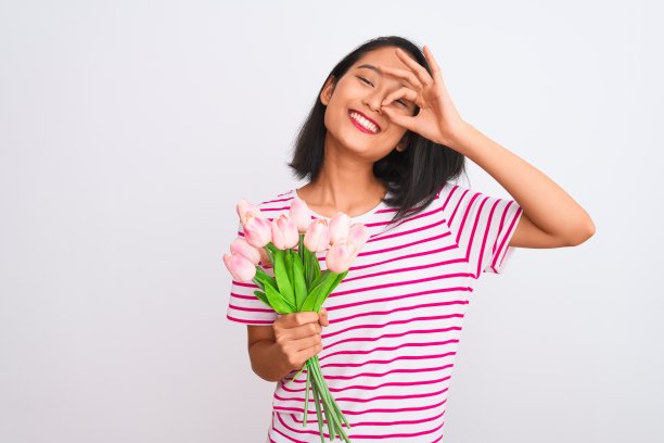 年轻的中国女子拿着玫瑰花束站在孤立的白色背景与快乐的脸微笑做ok手势与手在眼睛透过手指图片下载