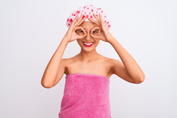 一个年轻的中国女人，穿着浴巾和浴帽，在孤立的白色背景下做着不错的手势，就像望远镜伸出舌头，眼睛透过手指看。疯狂的表情。图片下载