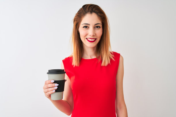 年轻漂亮的红发女人在白色背景下手拿咖啡，面带微笑图片下载