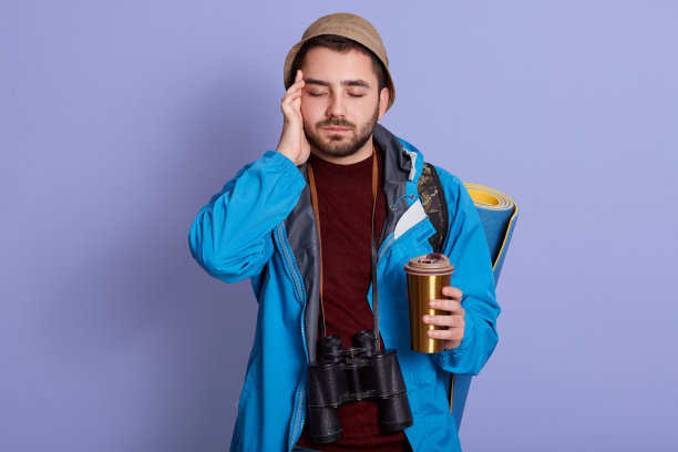 演播室照片：男子背着背包，以淡紫色为背景，拿着装有咖啡或茶的保温杯，手放在额头上，头痛，闭着眼睛站着。旅行，健康概念。图片下载