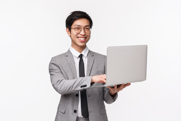 这是一个穿着灰色西装、站在白色背景下、手持笔记本电脑、微笑着对着镜头、远程工作、用电脑演示演示的自信成功的年轻亚洲男子的照片图片下载