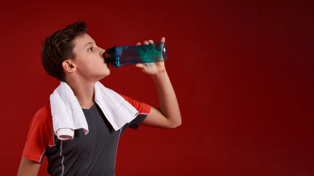 尽一切努力。一个十几岁的男孩正在运动，他正在喝瓶子里的水。孤立在红色背景上。健身、训练、积极的生活方式理念。水平。图片下载