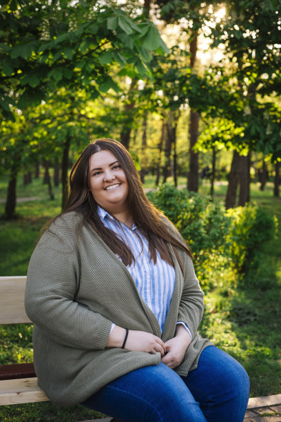 一位身穿条纹衬衫、绿色开衫和蓝色牛仔裤，坐在公园长椅上的快乐微笑的加大码女人图片下载