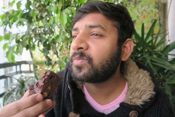 英俊的年轻印度男子拿着、吃着/咬着新鲜出炉的自制巧克力布朗尼，准备吃，背景是模糊的花园，留着胡子的男子穿着带人造毛皮帽子的棕色连帽衫，家庭烘焙照片图片下载