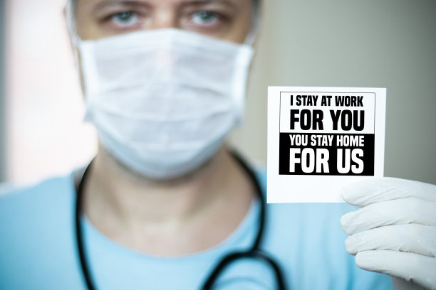 戴着口罩的绿眼中年医生拿着一张写着“请待在家里”的纸图片下载