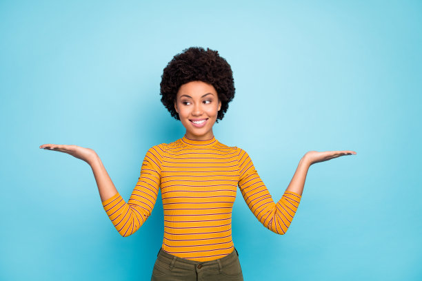 美丽的深色皮肤波浪女士的照片，打开手掌展示新奇的产品建议两个看起来最好的一个穿黄色条纹衬衫裤子孤立的蓝色背景图片下载