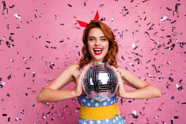 漂亮兴奋的女士的照片举行发光的迪斯科球主题复古派对跳舞明星五彩纸屑落下戴头巾点点连衣裙黄色皮带孤立的粉红色背景图片下载