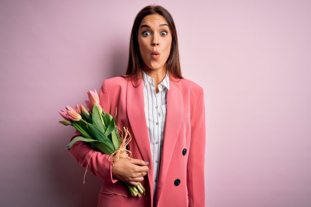 年轻美丽的黑发女子捧着一束郁金香花在粉红色的背景上吓了一跳，脸上带着惊讶，害怕和兴奋的恐惧表情图片下载