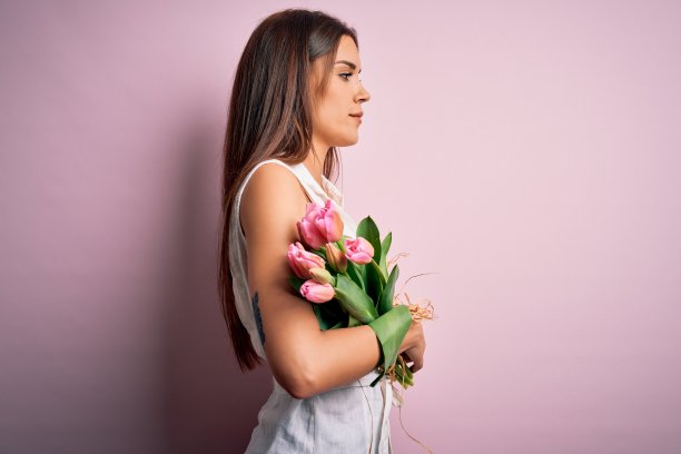 年轻美丽的黑发女子捧着郁金香花在粉红色的背景看向一边，放松的侧面姿势与自然的脸和自信的微笑。图片下载