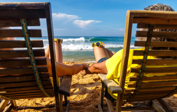 在阳光明媚的日子里，一对中年夫妇手牵着手，躺在木制日光浴床上欣赏海洋和云彩的景色图片下载