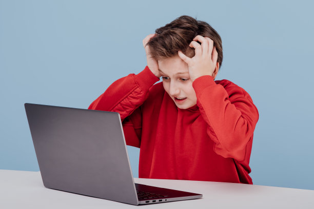 一个穿着红色运动衫，双手抱头的吓坏了的小男孩。在工作室的笔记本电脑旁边，蓝色背景图片下载