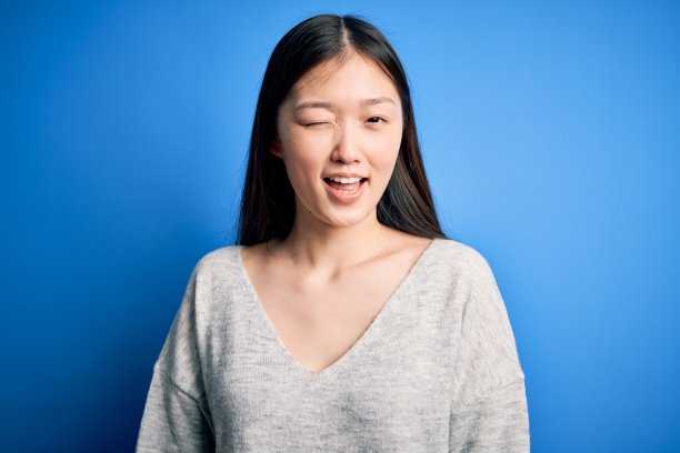 年轻美丽的亚洲女人穿着休闲毛衣站在蓝色孤立的背景眨眼睛看着相机与性感的表情，开朗和幸福的脸。图片下载