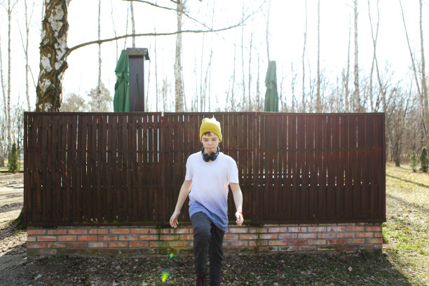 波兰阳光明媚的公园里，戴着黄色帽子和耳机，走在棕色栅栏前可爱的少年男孩的广角照片图片下载