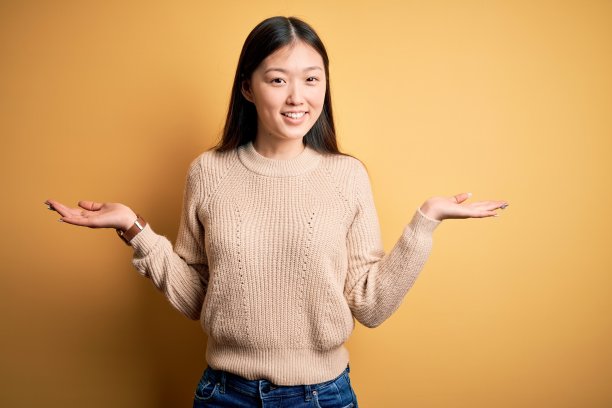年轻美丽的亚洲女人穿着休闲毛衣在黄色孤立的背景微笑显示双手张开的手掌，呈现和广告比较和平衡图片下载
