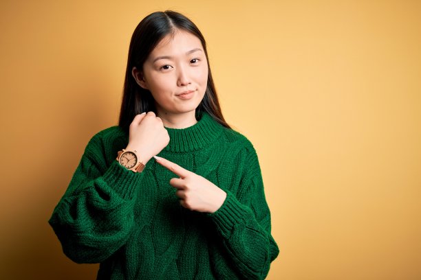 年轻美丽的亚洲女子穿着绿色的冬季毛衣越过黄色孤立的背景，匆忙地指着看着时间，不耐烦，看着镜头用轻松的表情图片下载