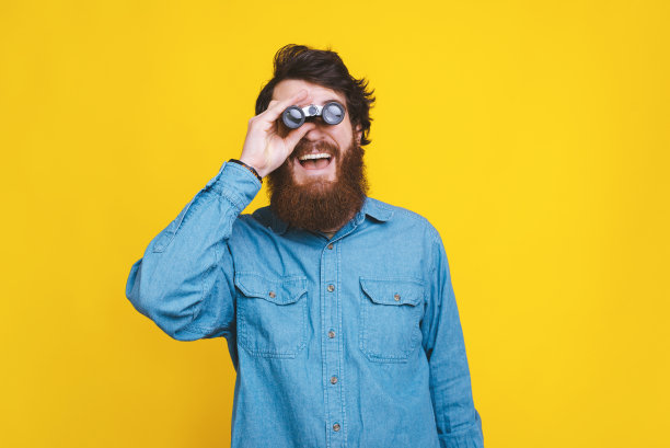 快乐的胡须男在黄色背景上手持双筒望远镜图片下载