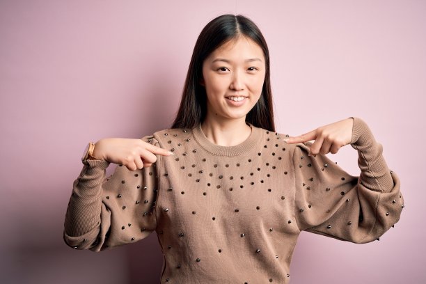 年轻美丽的亚洲女人穿着时尚和优雅的毛衣在粉红色孤独的背景看起来自信与微笑的脸，用手指指着自己骄傲和幸福。图片下载