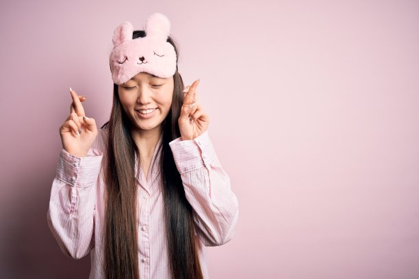 年轻的亚洲妇女穿着睡衣和睡眠面具在粉红色孤立的背景手势交叉手指微笑希望和闭上眼睛。运气和迷信的概念。图片下载