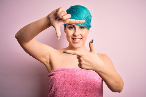 年轻女子与蓝色时尚头发穿着毛巾淋浴后在粉红色的背景微笑与手和手指框架与快乐的脸。创意和摄影概念。图片下载