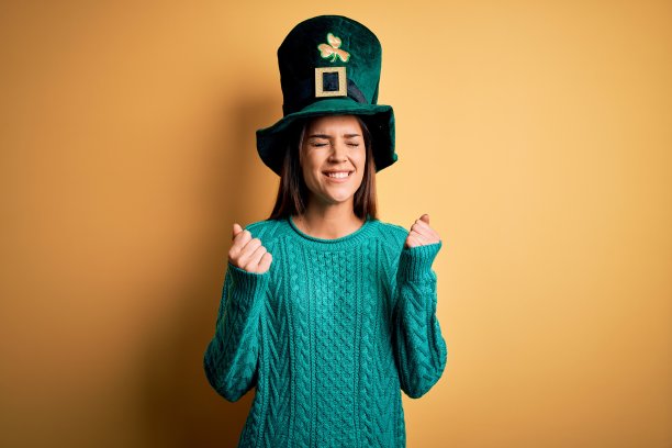 年轻美丽的黑发女人戴着绿帽子与三叶草庆祝圣帕特里克节兴奋的成功与双臂举起和眼睛闭上庆祝胜利微笑。赢家的概念。图片下载