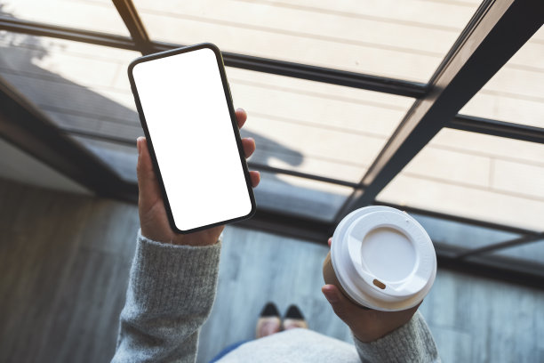 一个女人拿着一个黑色的手机和空白的白色桌面屏幕和咖啡杯图片下载
