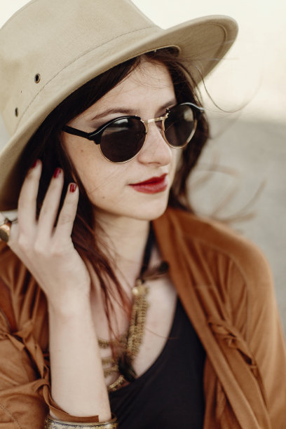 戴着太阳镜和帽子，穿着流苏披肩和皮包的时尚波西米亚风美女。在山区河流沙滩附近摆姿势的年轻吉普赛旅行者时髦女孩。性感的外表。图片下载