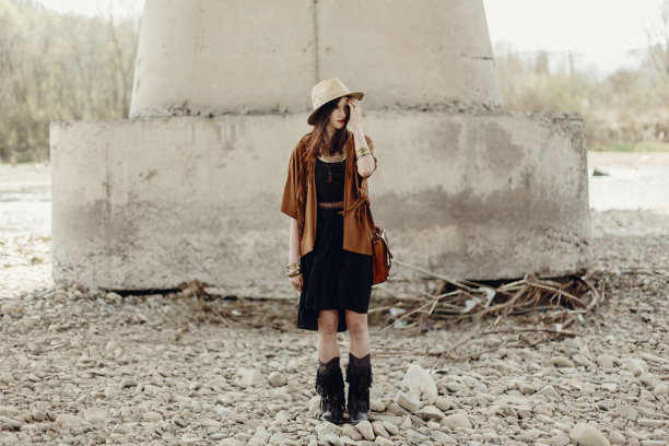 戴帽子、穿流苏披肩和靴子的时尚潮女在河边石头附近摆姿势，波西米亚风旅行，夏季旅行图片下载