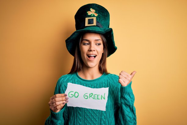 年轻美丽的女人戴着绿帽子庆祝圣帕特里克节，拿着信息指向和显示拇指向上到一边与幸福的脸微笑图片下载