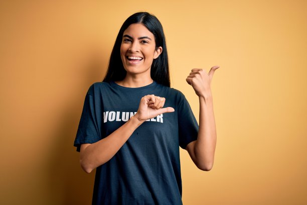 年轻美丽的西班牙妇女穿着志愿者t恤作为社会慈善道德指向背后的手和拇指，微笑自信图片下载