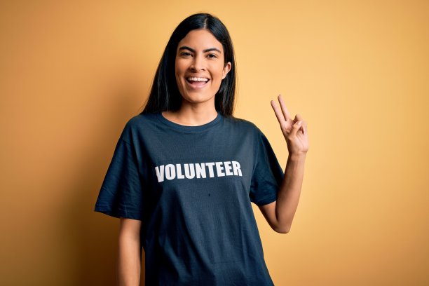 年轻美丽的西班牙妇女穿着志愿者t恤作为社会慈善道德微笑看镜头显示手指做胜利手势。二号人物。图片下载