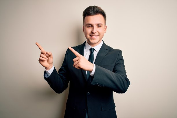 年轻英俊的商人穿着优雅的西装和领带在孤立的背景微笑，看着相机指向与双手和手指的一边。图片下载