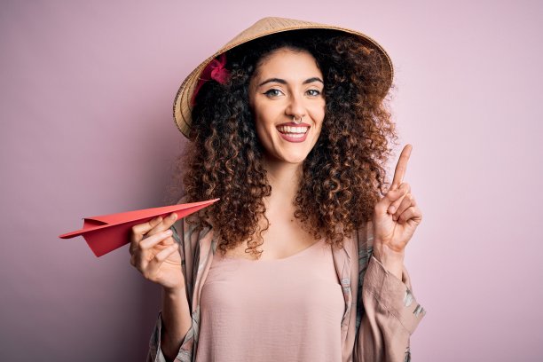 年轻漂亮的女人卷发和穿洞戴亚洲帽拿着纸飞机惊讶的想法或问题指着幸福的脸，第一图片下载