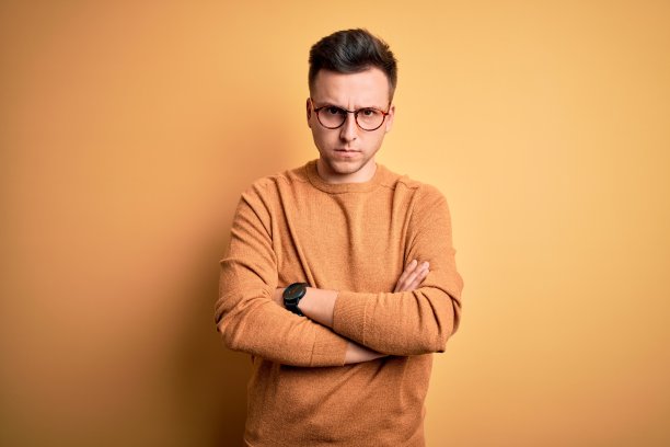 年轻英俊的白人男子戴眼镜和休闲冬季毛衣在黄色背景怀疑和紧张，不赞成的表情与交叉的胳膊。消极的人。图片下载