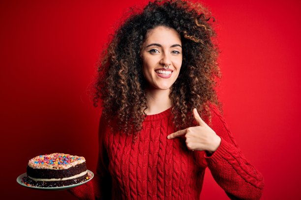 年轻美丽的女人与卷发和穿孔持有盘子与生日蛋糕与惊讶的脸指向自己的手指图片下载