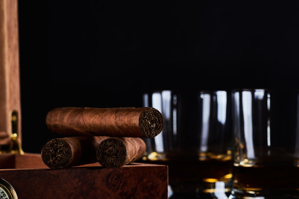 三支古巴雪茄，两杯威士忌或朗姆酒和带有湿度计和黑色背景的木箱的静物画。为文本留出空间。图片下载