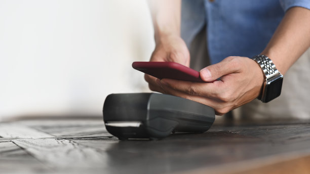 裁剪的图像，一个聪明的男人的手拿着智能手机，并使用NFC技术在信用卡读卡器上进行支付放置在咖啡馆付款柜台。技术和支付概念。图片下载