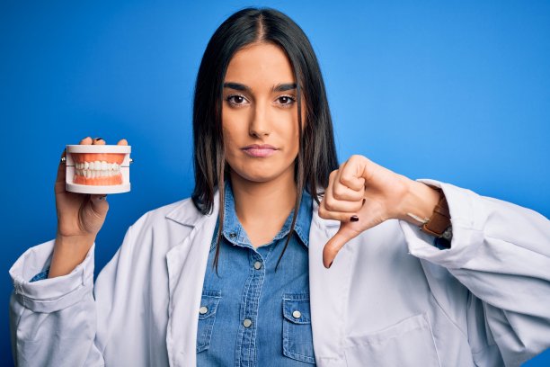 年轻美丽的黑发牙医女人穿着外套拿着塑料牙齿修复与愤怒的脸，消极的迹象显示不喜欢拇指下来，拒绝的概念图片下载
