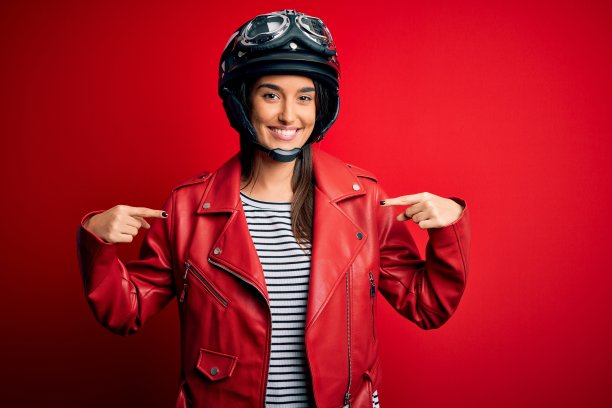 年轻美丽的黑发摩托车女戴着摩托车头盔和红色夹克看起来自信，脸上带着微笑，用手指指着自己骄傲和快乐。图片下载