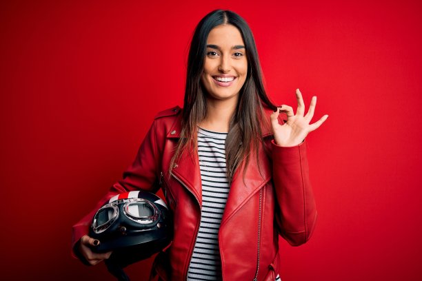 年轻漂亮的黑发摩托车女手持摩托车头盔和红色夹克做ok手势与手指，优秀的象征图片下载