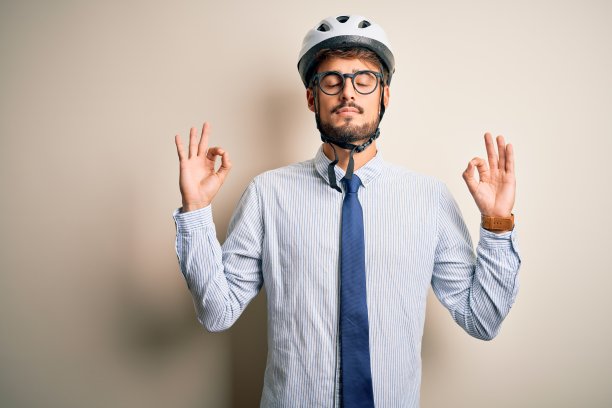 年轻的商人戴着眼镜和自行车头盔站在孤立的白色背景放松和微笑，闭上眼睛做冥想手势与手指。瑜伽的概念。图片下载