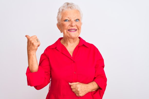 面带微笑的年长白人妇女站在白色背景前，面带笑容，翘起大拇指指向侧面图片下载
