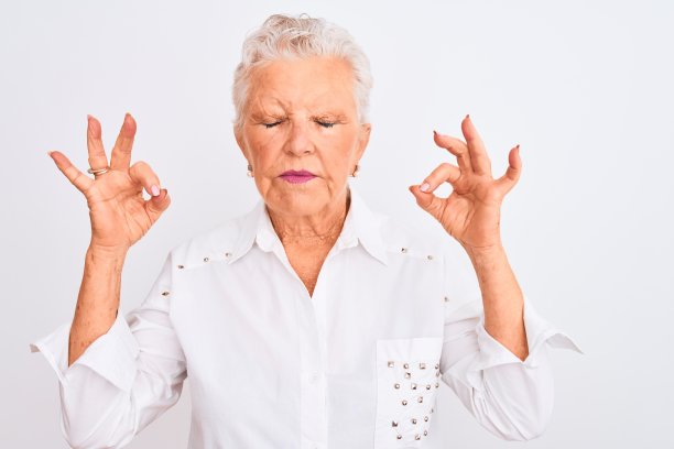 年长的灰发女人穿着优雅的衬衫站在白色的背景前，放松地闭着眼睛，用手指做冥想手势，面带微笑。瑜伽概念。图片下载