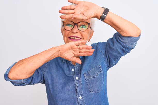 年长的白人妇女穿着牛仔衬衫和眼镜站在白色背景前，笑着，用手捂住脸玩躲猫猫，惊喜又兴奋图片下载