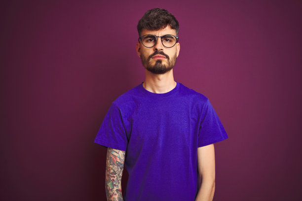 有纹身的年轻男人穿着T恤和眼镜，在紫色背景上表情严肃地看着镜头图片下载