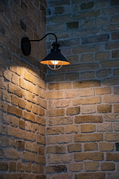 旧金属街灯在建筑物黑暗的角落里发光。风化的石头砖墙和金属灯罩与闪亮的灯泡。古老的砖砌图案背景与复制空间。图片下载