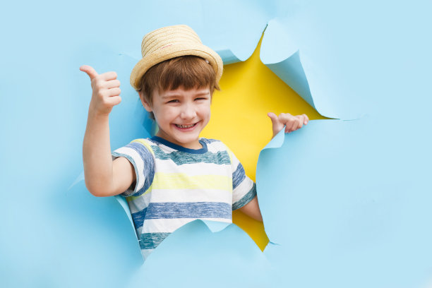 可爱的小男孩戴着一顶帽子，从撕破的纸洞里伸出大拇指摆姿势。纸张撕裂的效果。旅游和夏季休息的概念。图片下载