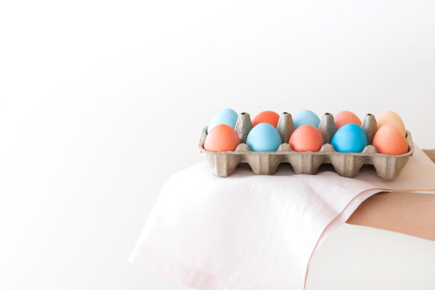 女人的手拿着纸盒上的粉色餐巾上画着红色和蓝色的复活节彩蛋近。逾越节的概念。白色背景。图片下载